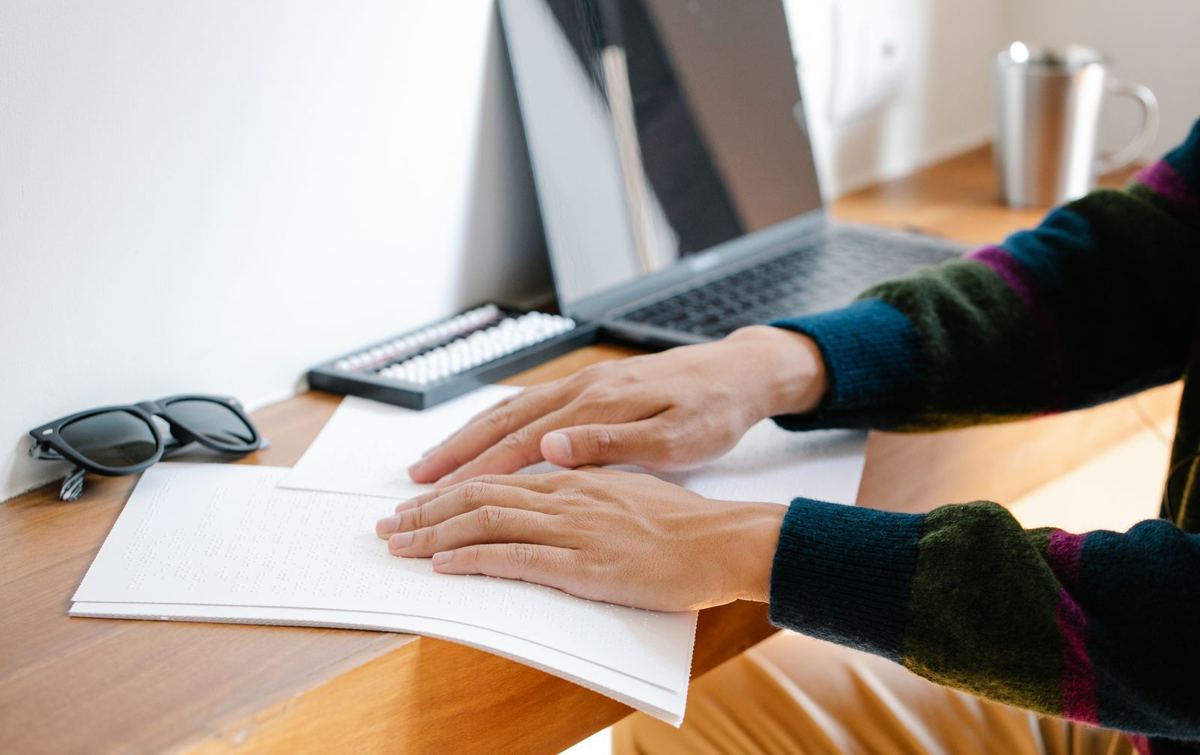 Two hands resting on a page printed in braille, in front of a computer keyboard