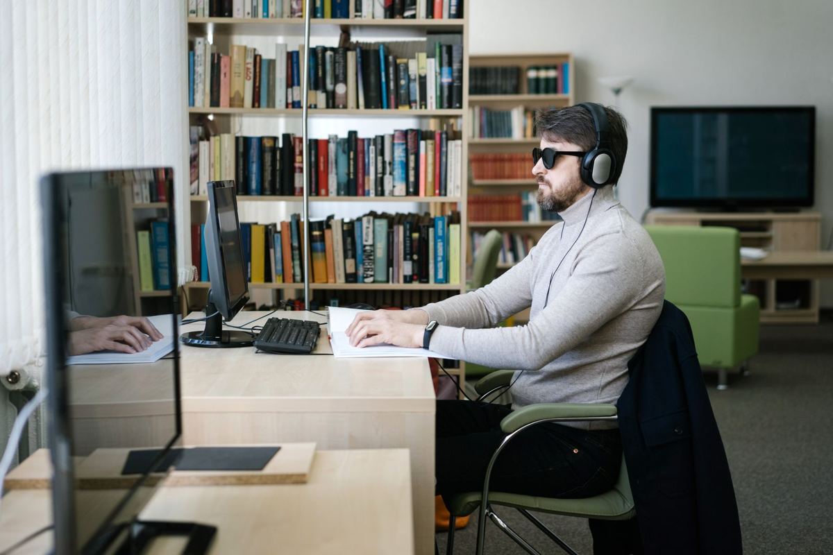 A blind man wearing dark glasses works on a computer in a library